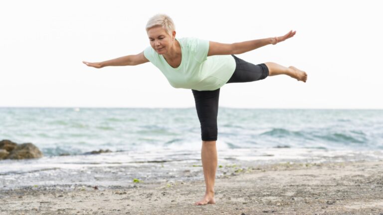Eine Frau in einem hellgrünen Shirt und schwarzen Leggings balanciert barfuß am Strand, mit Blick auf das Wasser.