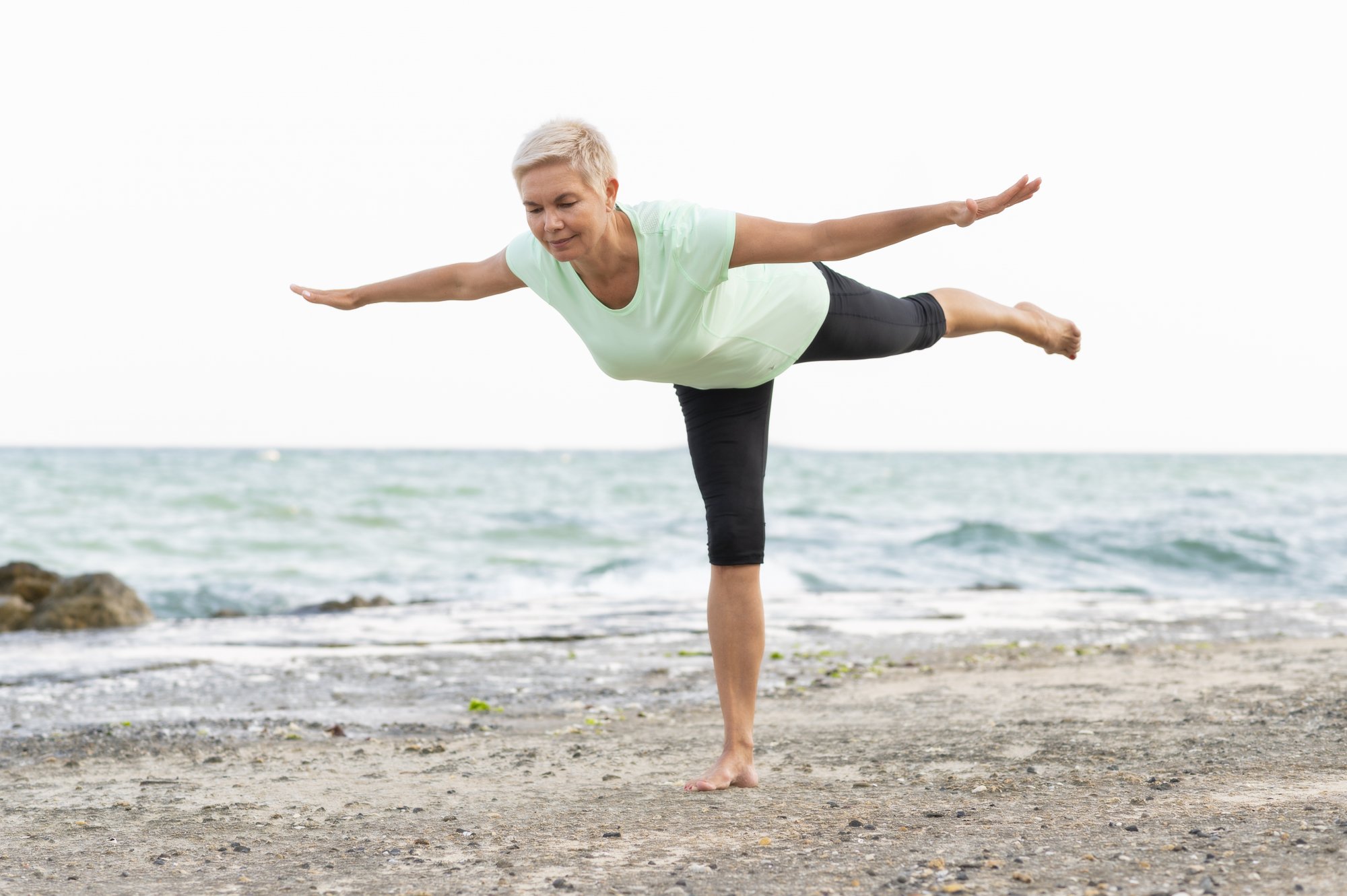 Balance_Training.jpg Eine Frau in einem hellgrünen Shirt und schwarzen Leggings balanciert barfuß am Strand, mit Blick auf das Wasser.