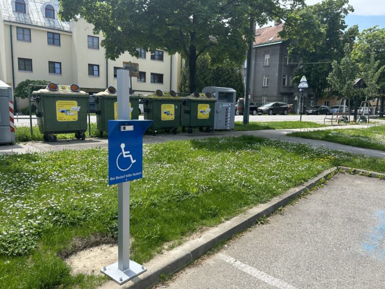 Blaues Schild mit dem Symbol für Barrierefreiheit und dem Hinweis 'Bei Bedarf bitte läuten' an einem Müllplatz.