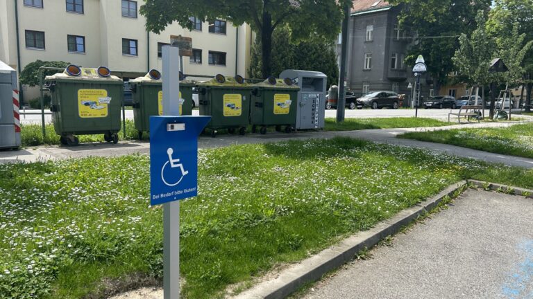 Blaues Schild mit dem Symbol für Barrierefreiheit und dem Hinweis 'Bei Bedarf bitte läuten' an einem Müllplatz.