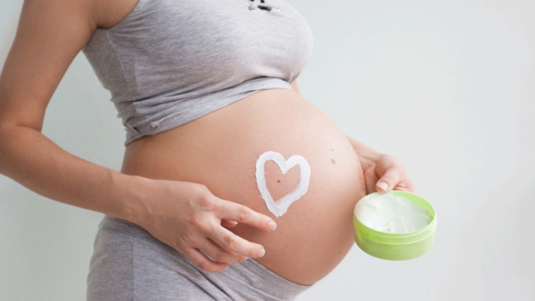 Eine schwangere Person trägt eine Crememaskierung in Herzform auf ihren Bauch auf, während sie eine grüne Dose hält.