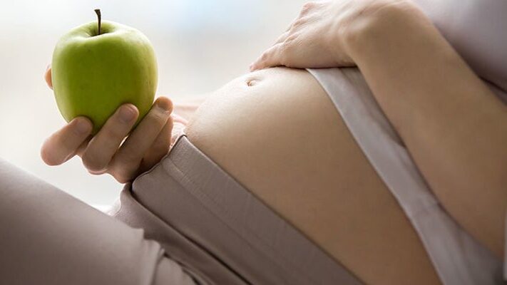 Eine schwangere Person hält einen grünen Apfel und berührt sanft ihren Bauch.