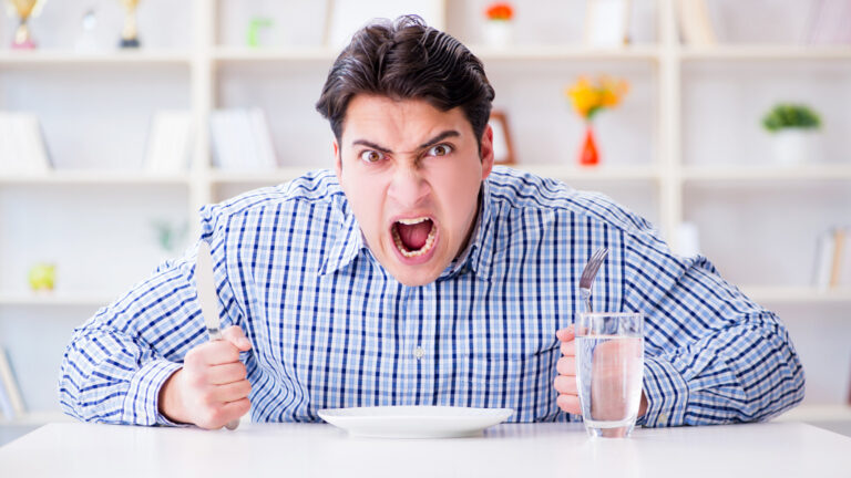 Ein wütender Mann hält ein Messer und eine Gabel über einem leeren Teller, während er an einem Tisch sitzt.