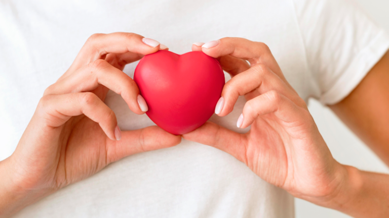 Eine Person hält ein rotes Herzen in beiden Händen vor einem hellen Hintergrund.