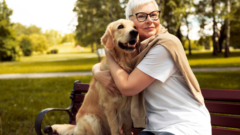 Eine Frau mit kurzen, blonden Haaren und Brille umarmt einen golden retriever auf einer Bank im Park.