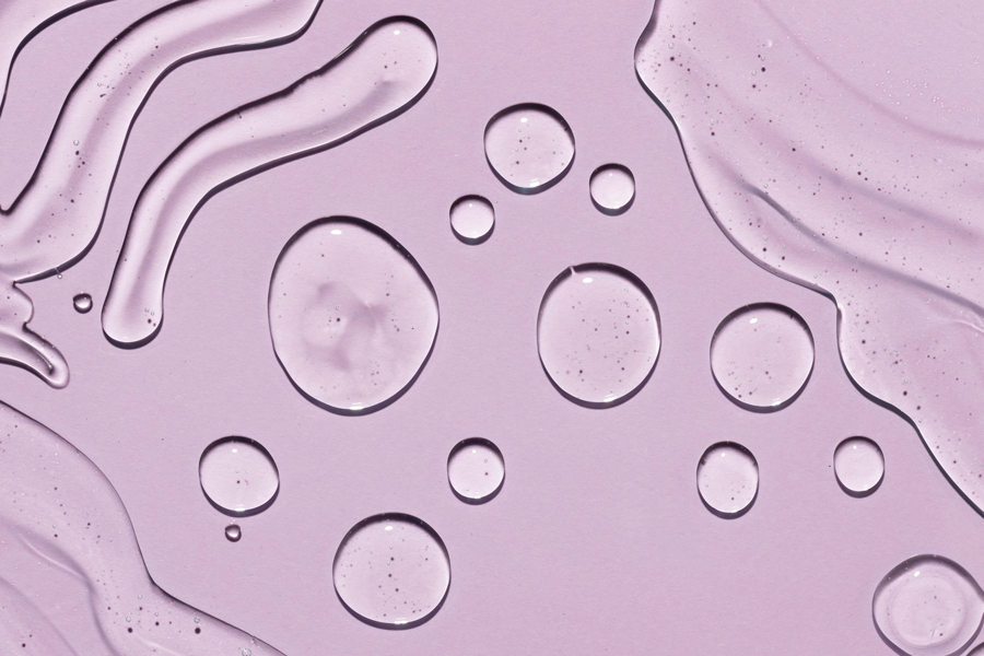 Hyaluron-web.png Transparente Wassertropfen auf einem helllila Hintergrund, mit fließenden Linien und Blasen.