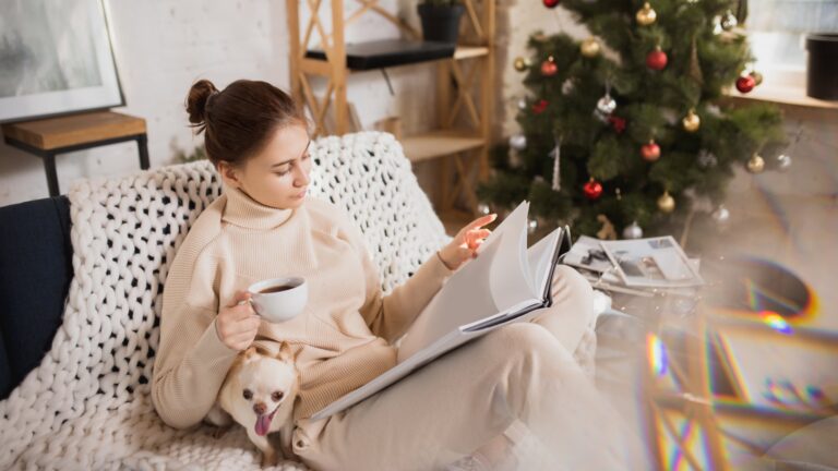 Eine Frau in bequemer Kleidung sitzt auf einem Sofa, liest in einem Buch und hält eine Tasse Kaffee, während ein Hund bei ihr sitzt.