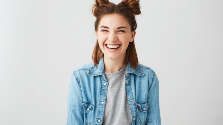 Lachende junge Frau mit Duttfrisur, trägt eine Jeansjacke über einem grauen T-Shirt, vor einem hellen Hintergrund.