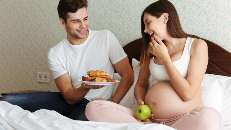 Ein schwangeres Paar sitzt im Bett, der Mann bietet der Frau mit einem Lächeln eine Platte mit Gebäck an, während sie einen Apfel hält.