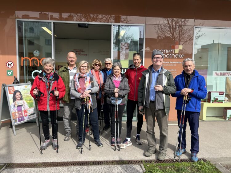 Gruppe von Personen mit Nordic-Walking-Stöcken vor einer Apotheke, lächelnd und in sportlicher Kleidung.
