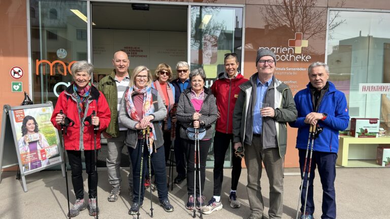Gruppe von Personen mit Nordic-Walking-Stöcken vor einer Apotheke, lächelnd und in sportlicher Kleidung.