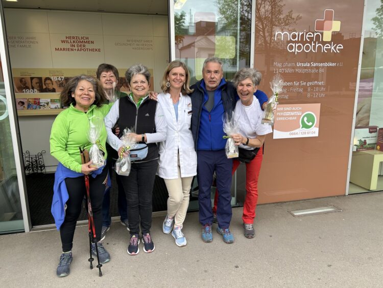 Gruppe von sechs Personen vor einer Apotheke, einige halten Tüten, fröhliche Atmosphäre, Apotheke im Hintergrund sichtbar.