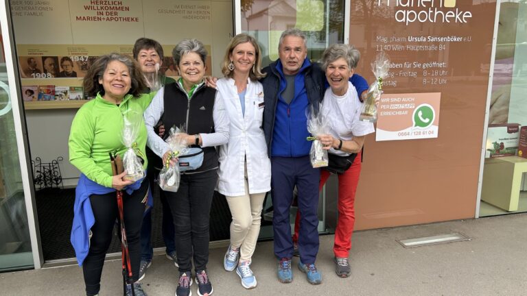 Gruppe von sechs Personen vor einer Apotheke, einige halten Tüten, fröhliche Atmosphäre, Apotheke im Hintergrund sichtbar.