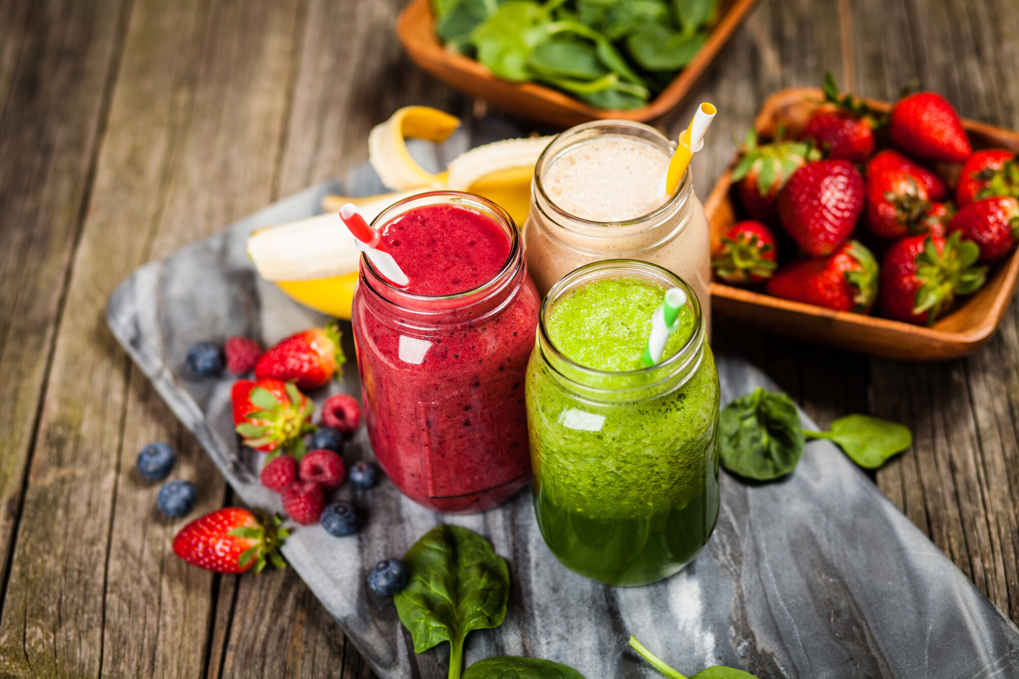 Drei bunte Smoothies in Gläsern mit Strohhalmen, umgeben von frischen Früchten und Spinat auf einer Holzunterlage.