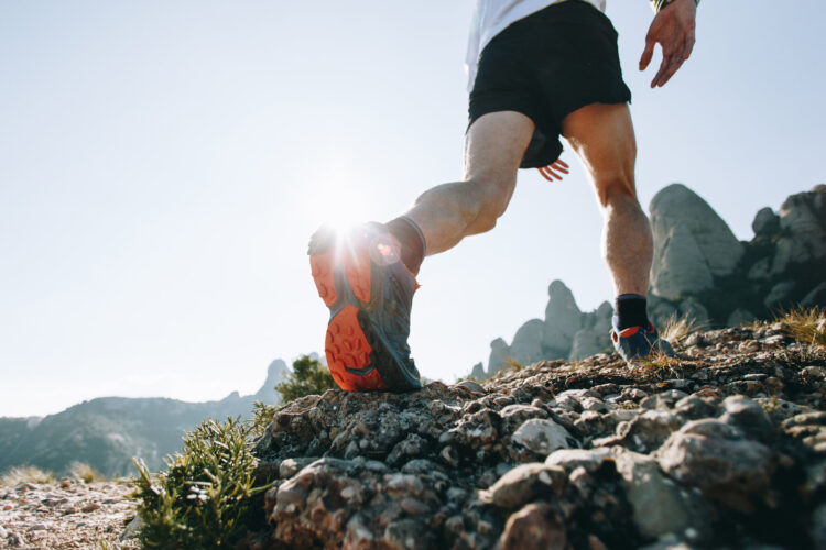 Läufer in sportlichen Schuhen auf einem kies- und steinigen Weg mit Berglandschaft im Hintergrund