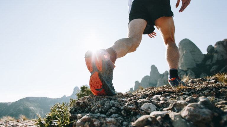 Läufer in sportlichen Schuhen auf einem kies- und steinigen Weg mit Berglandschaft im Hintergrund