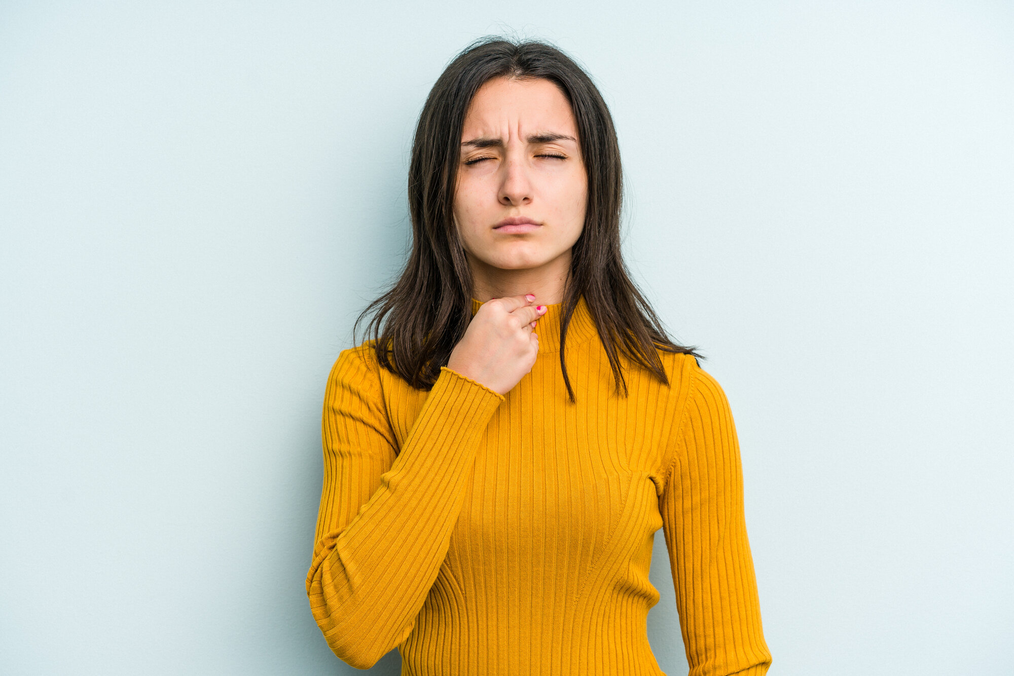 Stimme_versagt.jpg Frau in gelbem Pullover, die nachdenklich auf ihre Kehle zeigt, vor einer hellblauen Wand.