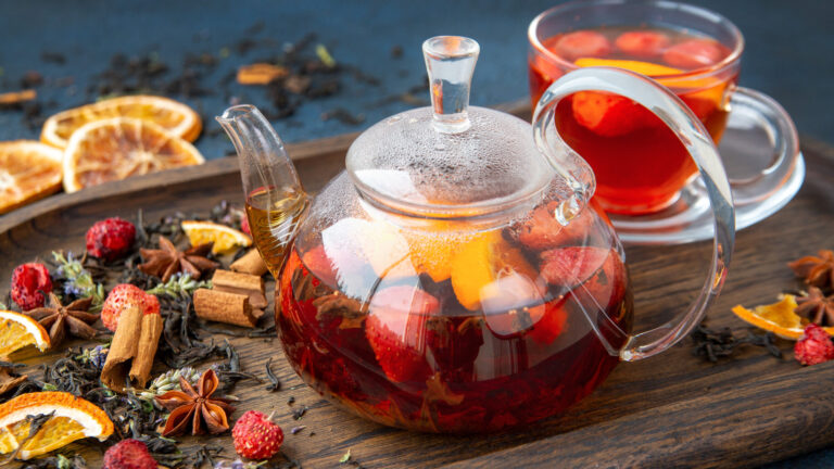 Teekanne aus Glas mit fruchtigem Tee, umgeben von Trockenfrüchten und Gewürzen auf einem Holzbrett.