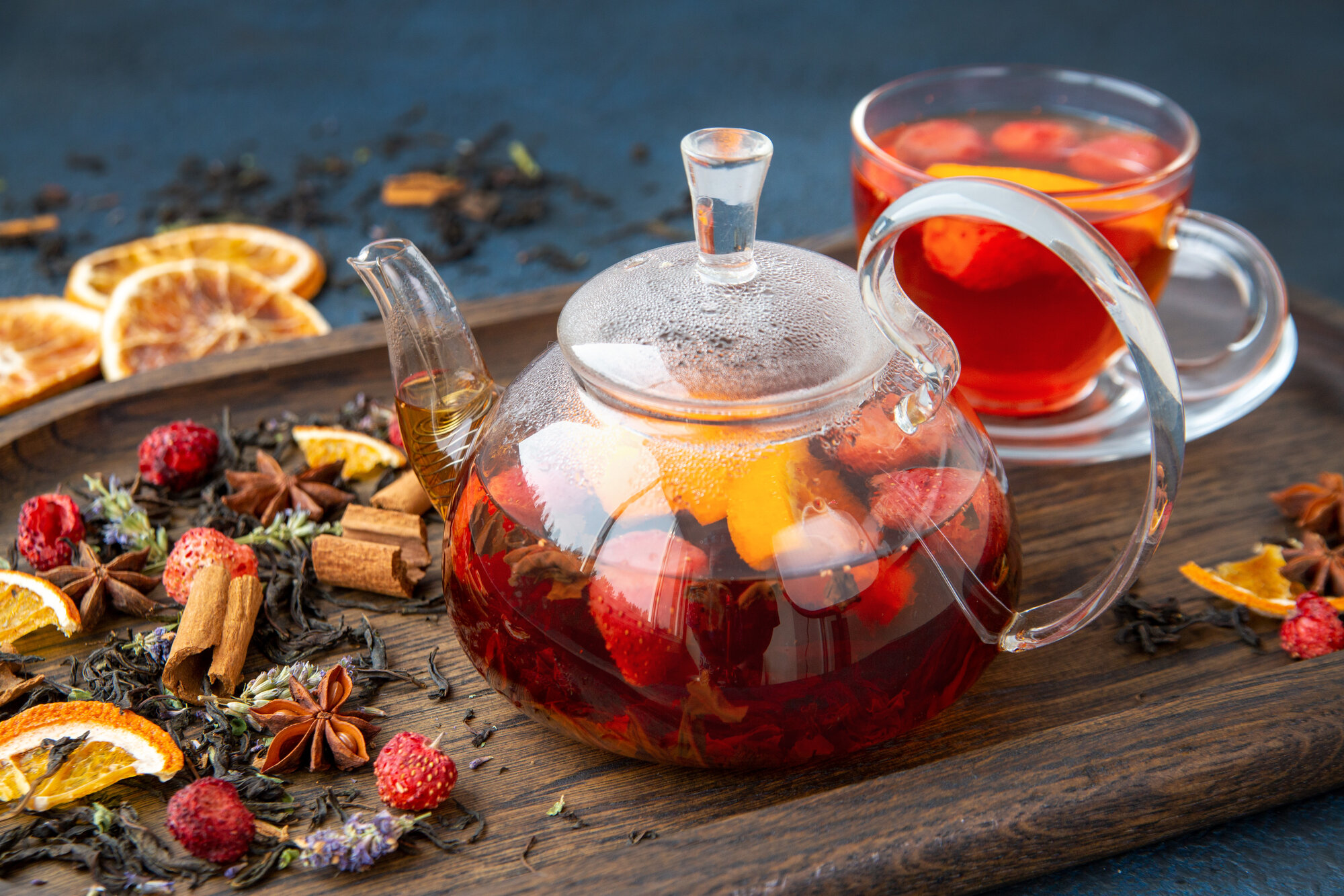 Teekanne aus Glas mit fruchtigem Tee, umgeben von Trockenfrüchten und Gewürzen auf einem Holzbrett.