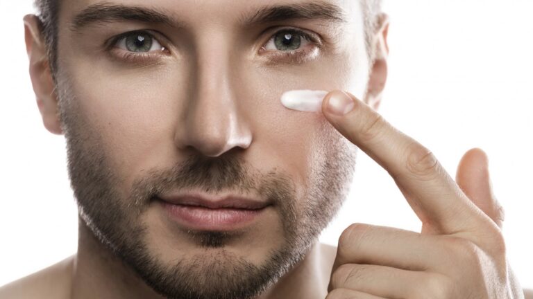 Ein Mann trägt eine Creme auf seine Wange auf, während er in die Kamera schaut. Hintergrund ist neutral.