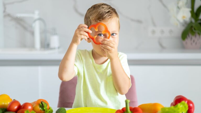 Ein Junge hält eine rote Paprika in Form eines Blumenmotivs und schaut in die Kamera, umgeben von frischem Gemüse.