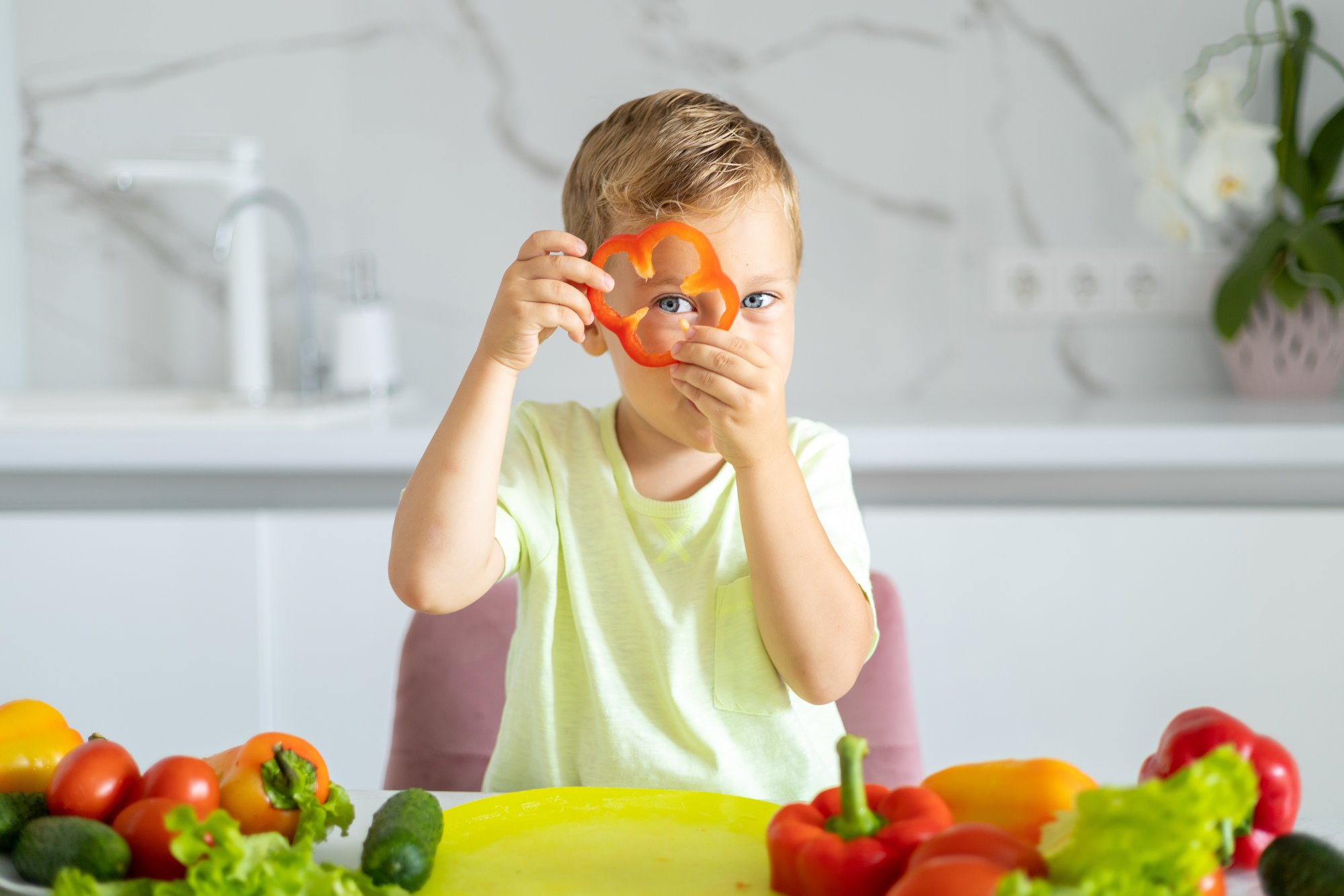 Vitaminmangel_erkennen.jpg Ein Junge hält eine rote Paprika in Form eines Blumenmotivs und schaut in die Kamera, umgeben von frischem Gemüse.