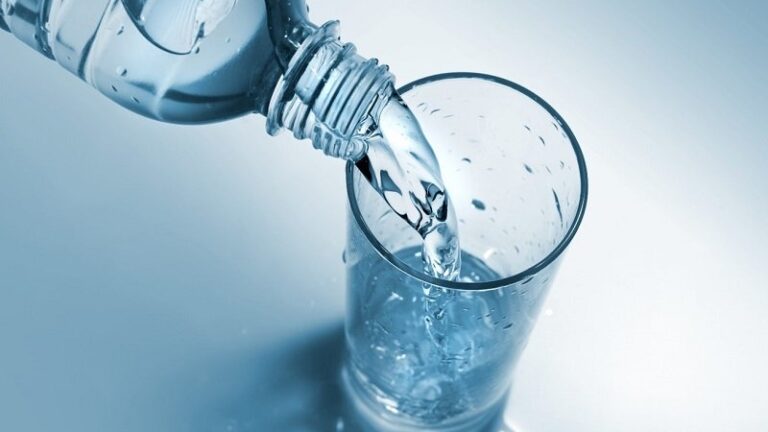 Eine Wasserflasche schüttet frisches Wasser in ein Glas, das auf einer hellen, blauen Oberfläche steht.