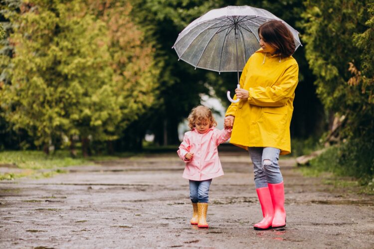 Wetterfuehligkeit.jpg Eine Frau in einem gelben Regenmantel hält einen Regenschirm, während sie mit einem kleinen Mädchen in einem rosa Regenmantel spaziert. Beide tragen Gummistiefel.