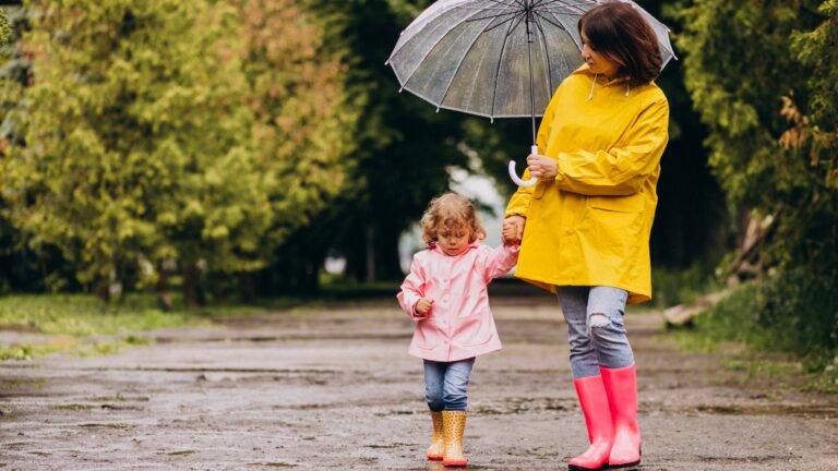 Eine Frau in einem gelben Regenmantel hält einen Regenschirm, während sie mit einem kleinen Mädchen in einem rosa Regenmantel spaziert. Beide tragen Gummistiefel.