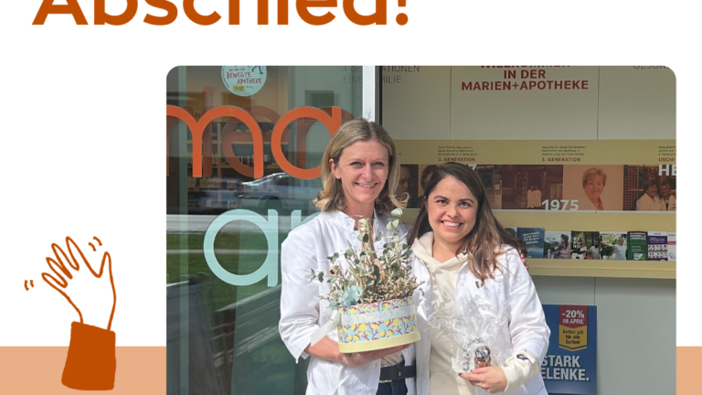 Zwei Frauen in weißen Kitteln stehen vor einer Apotheke und halten Geschenke, während sie sich verabschieden.