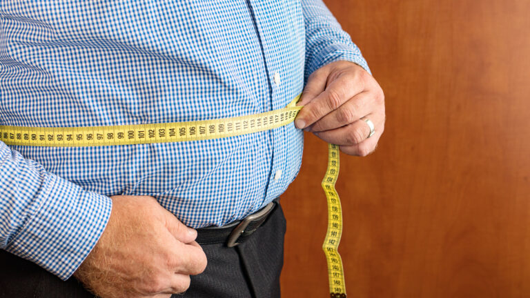 Mann misst seinen Bauchumfang mit einem gelben Maßband, trägt ein kariertes Hemd und dunkle Hose.