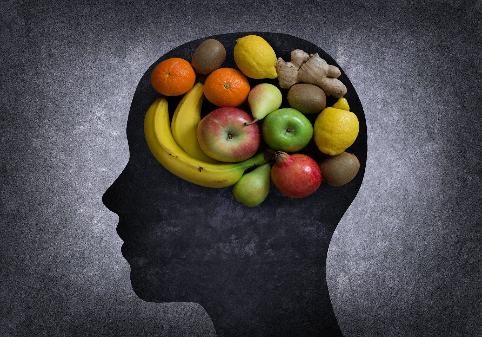 brainfood.jpg Silhouette eines Kopfes, gefüllt mit verschiedenen Früchten wie Äpfeln, Bananen, Orangen und Zitrusfrüchten auf dunklem Hintergrund.