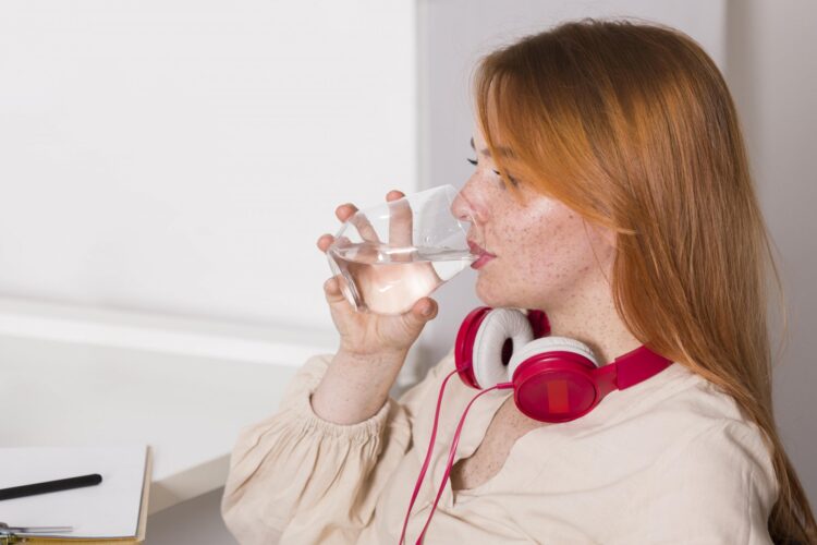 Eine Person mit roten Kopfhörern trinkt aus einem Glas Wasser und sitzt an einem Schreibtisch.