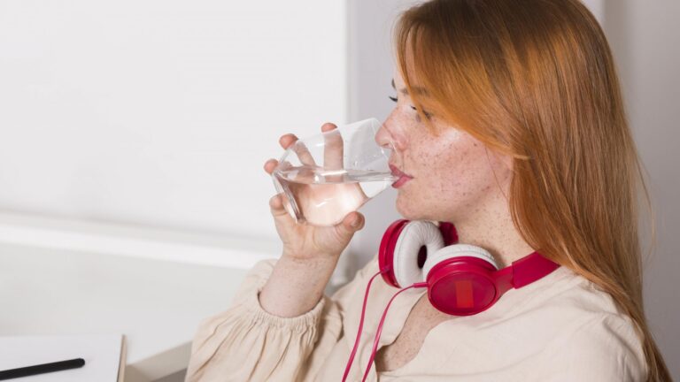 Eine Person mit roten Kopfhörern trinkt aus einem Glas Wasser und sitzt an einem Schreibtisch.