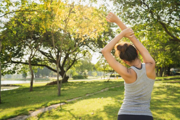 Eine Frau in einem grauen Tanktop dehnt sich in einem parkähnlichen Gelände mit Bäumen und Sonnenlicht.