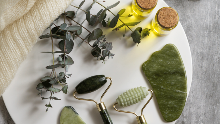 Ein weißer Teller mit drei Jade-Rollern, einem gua sha Stein, zwei Fläschchen ätherischem Öl und Eukalyptuszweigen.