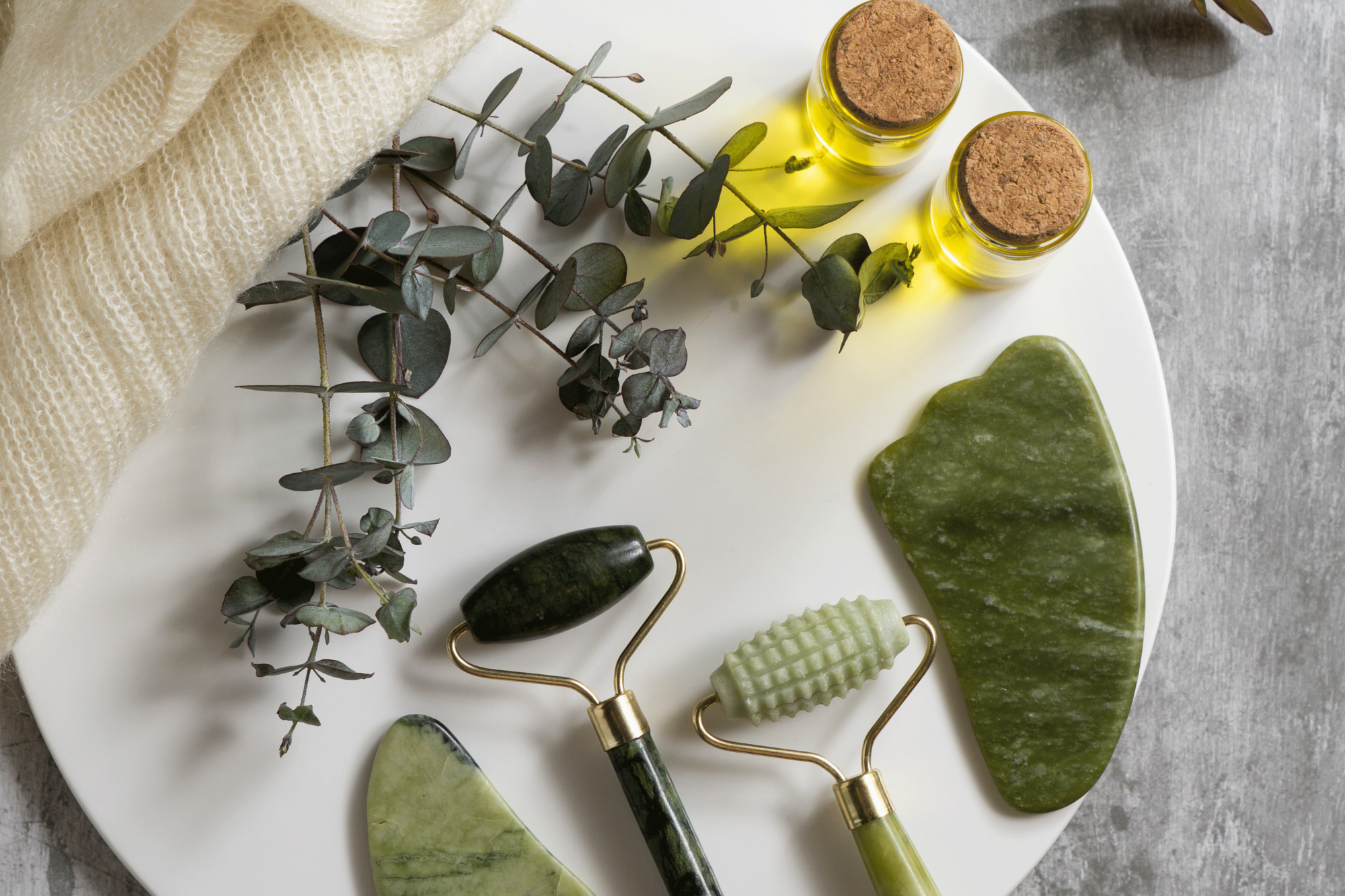 Ein weißer Teller mit drei Jade-Rollern, einem gua sha Stein, zwei Fläschchen ätherischem Öl und Eukalyptuszweigen.