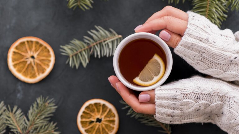 Eine Person hält eine Tasse mit Tee und einer Zitronenscheibe, umgeben von trockenen Orangenscheiben und Tannenzweigen.