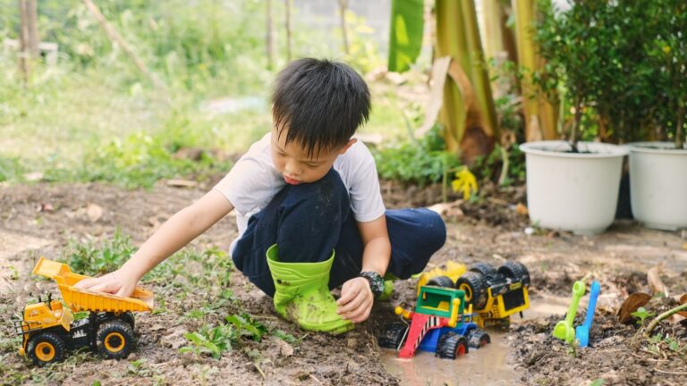 Ein Junge im Garten spielt mit verschiedenen Spielzeugfahrzeugen, darunter ein Lkw und ein Bagger, während er in matschigem Boden sitzt.