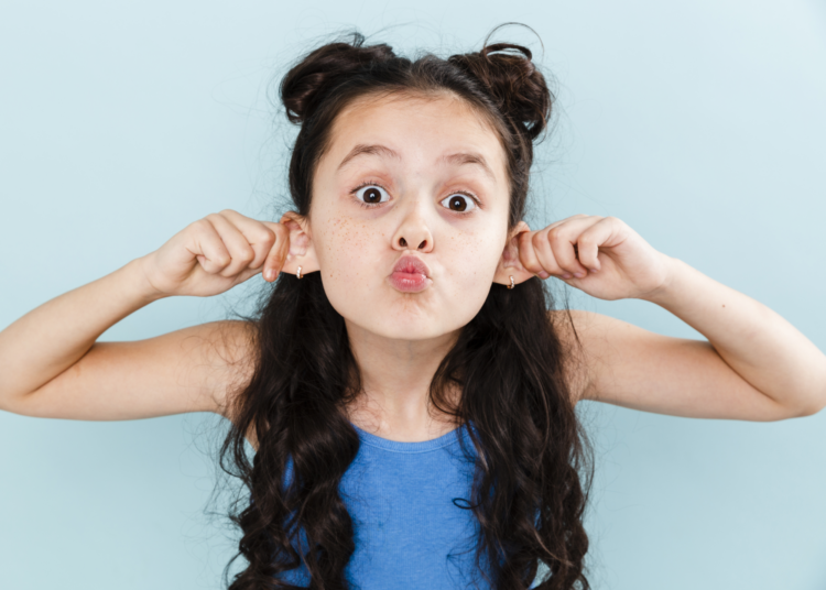 Ein Mädchen mit langen, lockigen Haaren zieht an ihren Ohren und macht ein lustiges Gesicht vor einer hellblauen Wand.