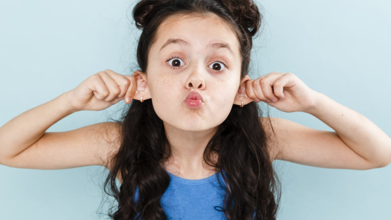 Ein Mädchen mit langen, lockigen Haaren zieht an ihren Ohren und macht ein lustiges Gesicht vor einer hellblauen Wand.