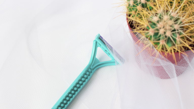 Ein grüner Rasierer liegt neben einem Topf mit einem Kaktus, umgeben von einem transparenten Stoff.