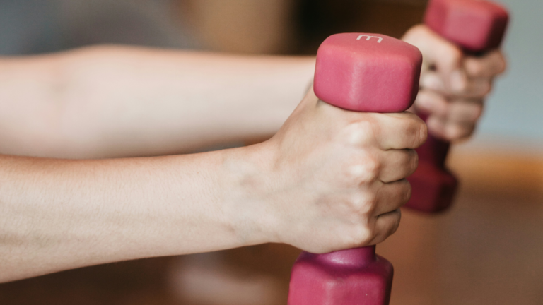 Hände halten zwei pinkfarbene Kurzhanteln in einem Fitnessstudio.