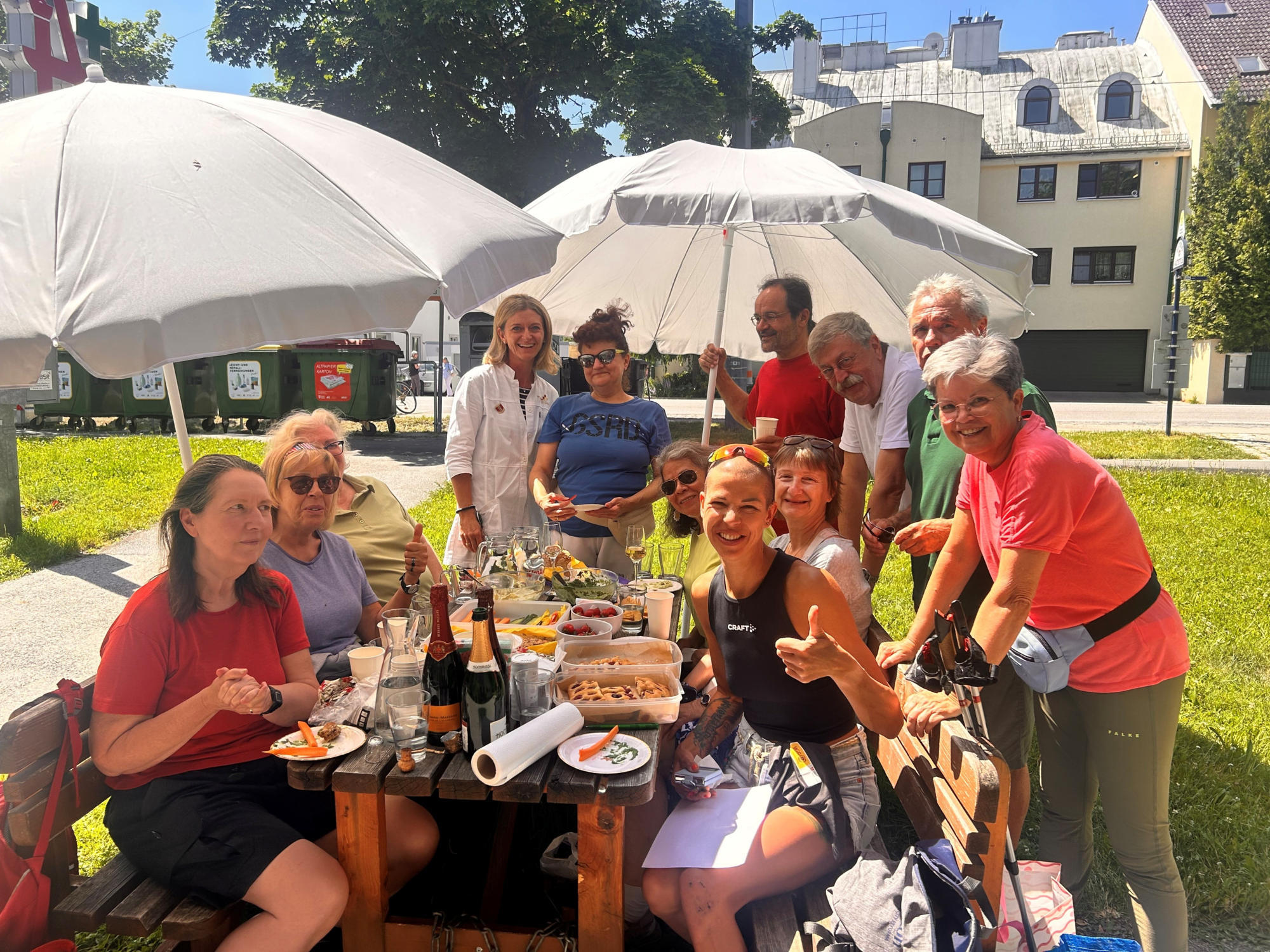 Nordic_Walking_Juli_2025.png Eine Gruppe von Menschen sitzt unter Sonnenschirmen an einem Tisch mit Speisen und Getränken im Freien.