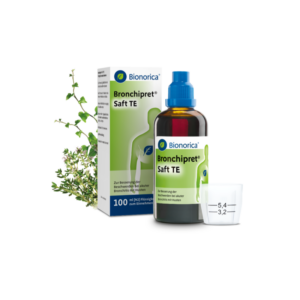 Bronchipret Saft TE in einer braunen Flasche, mit grüner Verpackung und einem Messbecher daneben, umgeben von Kräutern.