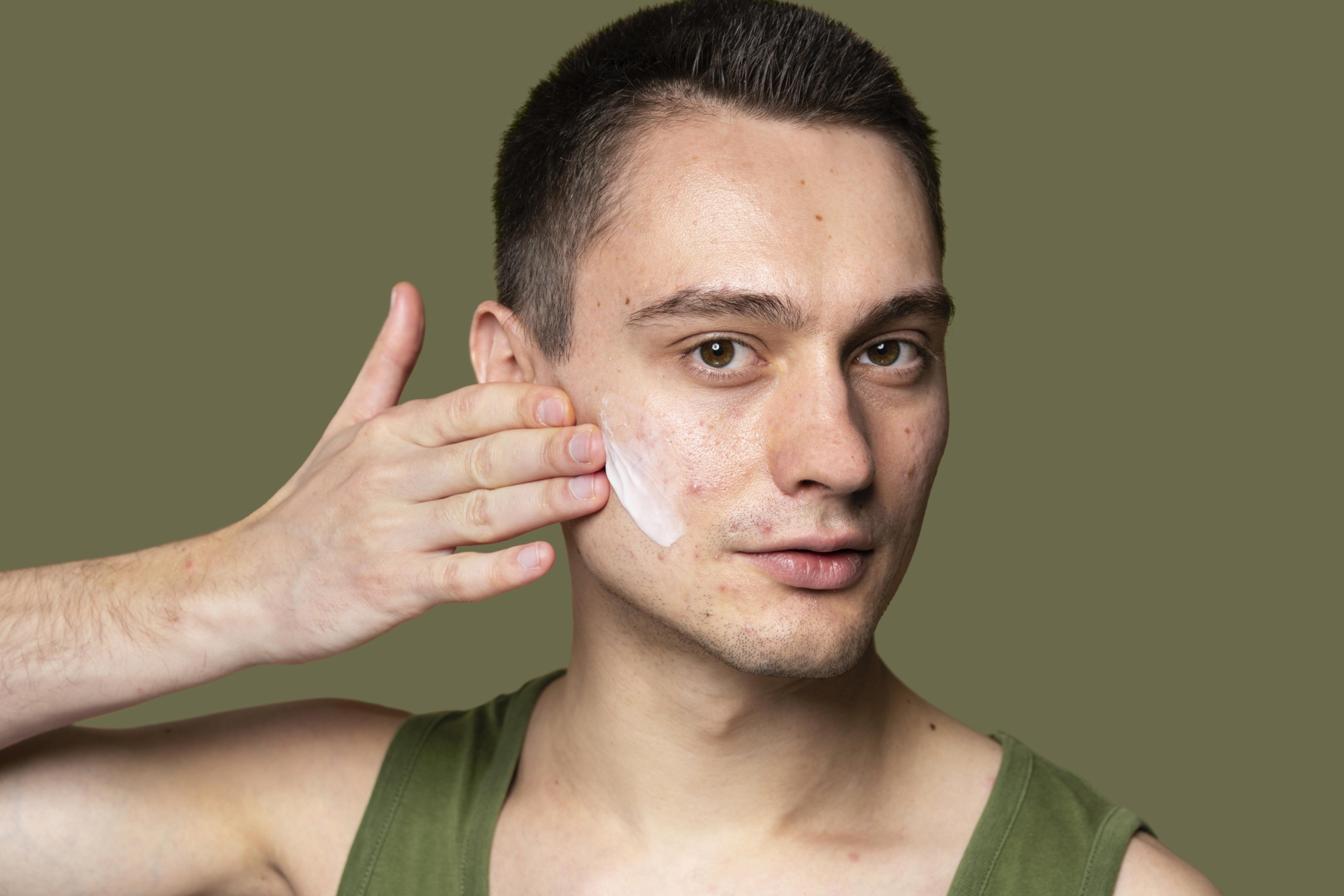 Unreine_Haut.png Ein Mann trägt Gesichtscreme auf seine linke Wange auf, Hintergrund ist ein einfarbiger, olivgrüner Farbton.