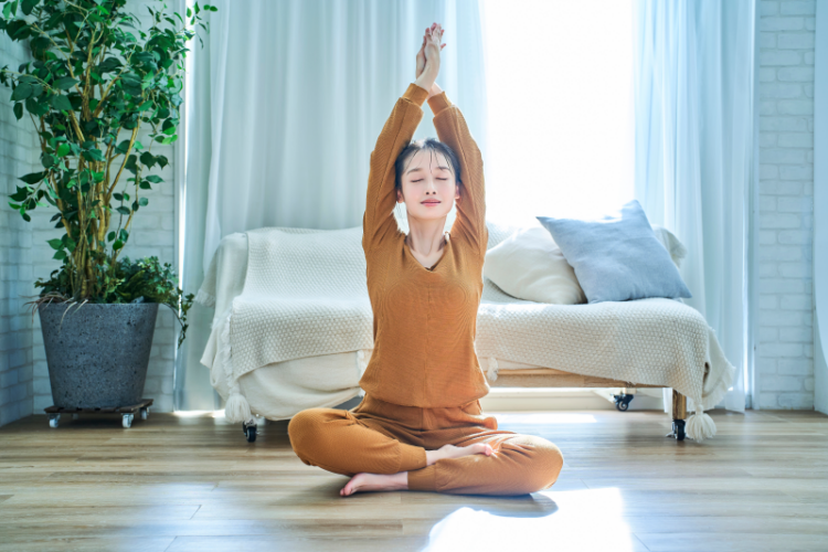 Eine Person in orangefarbenem Yoga-Outfit sitzt im Schneidersitz und streckt die Arme nach oben, umgeben von heller, luftiger Wohnungseinrichtung.