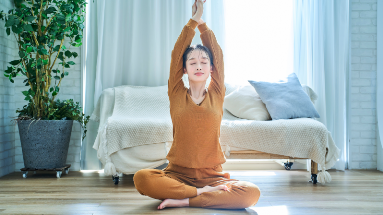 Eine Person in orangefarbenem Yoga-Outfit sitzt im Schneidersitz und streckt die Arme nach oben, umgeben von heller, luftiger Wohnungseinrichtung.