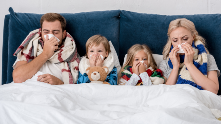 Eine Familie mit zwei Kindern kuschelt sich in Decken auf einer Couch, alle halten Taschentücher vor den Nasen.