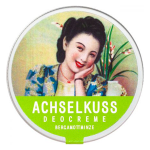 Runde Verpackung einer Deocreme mit dem Namen "Achselskuss" und Abbildung einer lächelnden Frau in traditioneller Kleidung.
